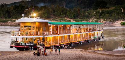Children stand in front of a wooden traditional cruise boat at dusk in the Laos portion of the Mekong River