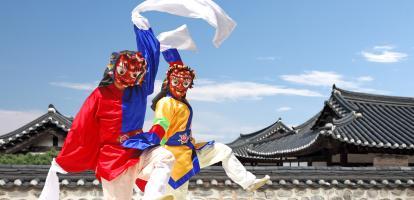 Two traditional folk dancers wearing masks dance in front of hanok houses in South Korea