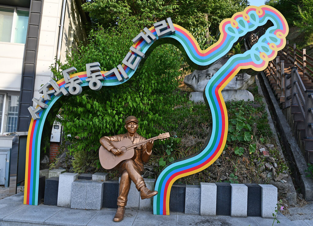 Brass statue of a man playing his guitar, sitting under a bigger guitar frame