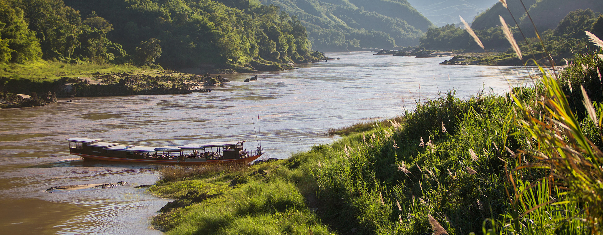 Laos-Pakbeng-Luang-Say-Lodge-and-Cruises-Hero Long, slim boat docking on a river next to forested hills in Pakbeng, Laos