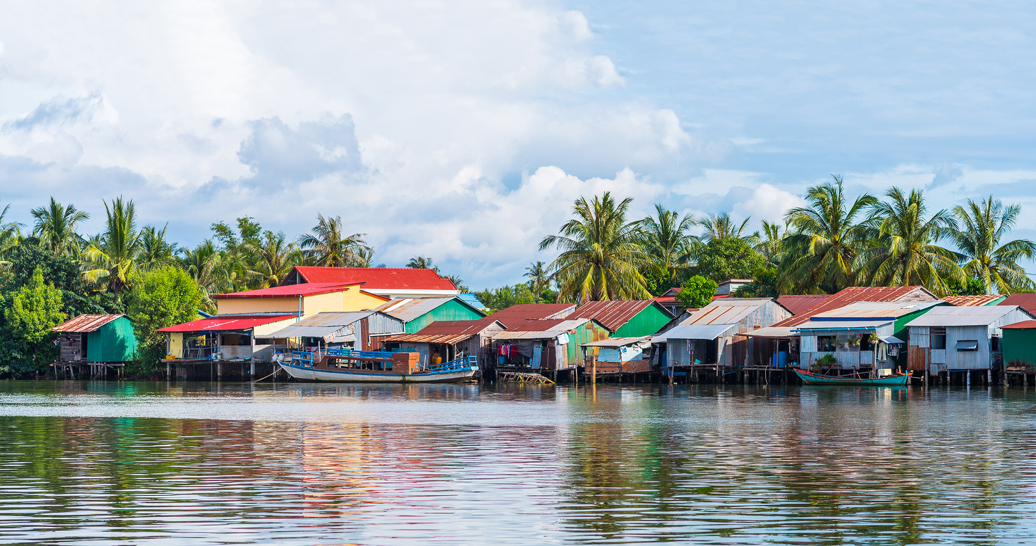 Kampot-floating-village-David-Bokuchava-Hero Riverside village has multi coloured houses built right on the river, with barges parked next to them and slightly clouded blue skies