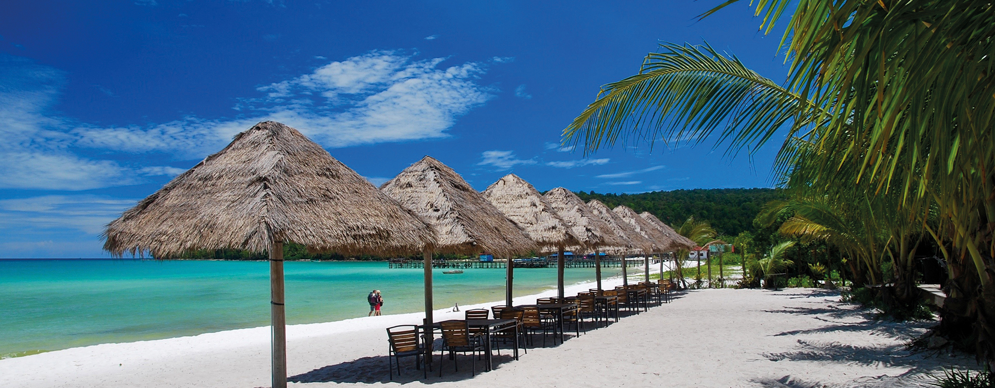 Koh_Rong_Beach_Hero_Cambodia_B White-sand beach lined with straw-thatched umbrellas next to azure waters in Cambodia's coast.