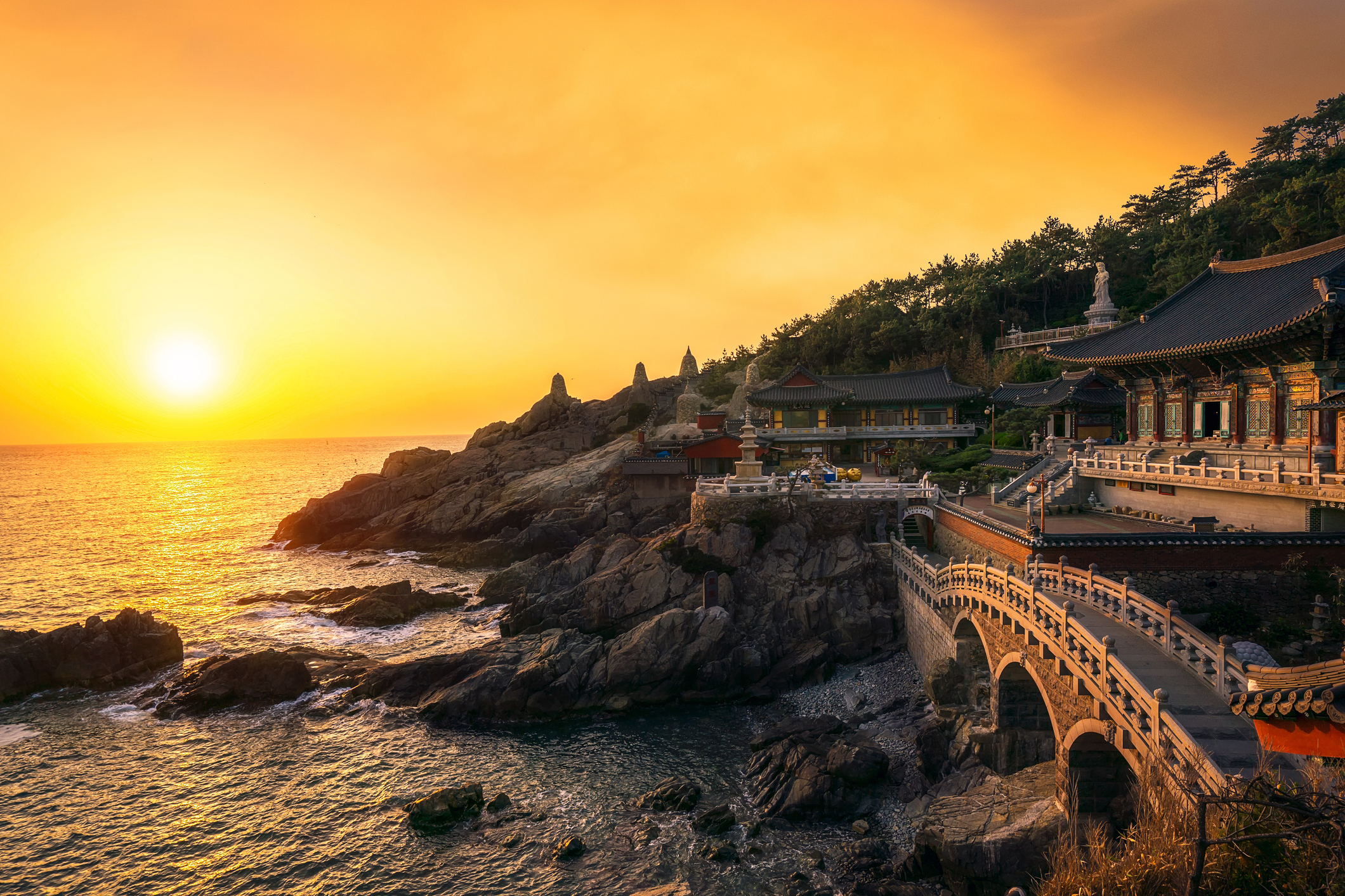 Yonggungsatemple-temple-in-Busan.jpg Sun setting over sea next to ornate temple and bridge in Busan