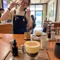 Close-up of Vietnamese egg coffee on a wooden table in a rustic café, with a cheerful barista posing in the background.