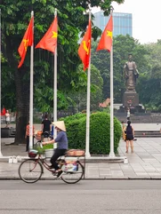 A person in a traditional hat cycling in front of six poles waving the Vietnamese flag with a famous statue in the background