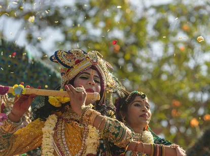 A person dressed in vibrant yellow and gold traditional attire, adorned with an ornate crown, jewelry, and flower garlands, plays a decorated flute while another person stands beside them in green attire and jewelry. Flower petals are falling around them, with a blurred background of trees and sunlight.