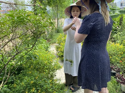Two people wearing Vietnamese conical hats walking through a tropical garden with green plants, yellow flowers, and banana trees on a sunny day