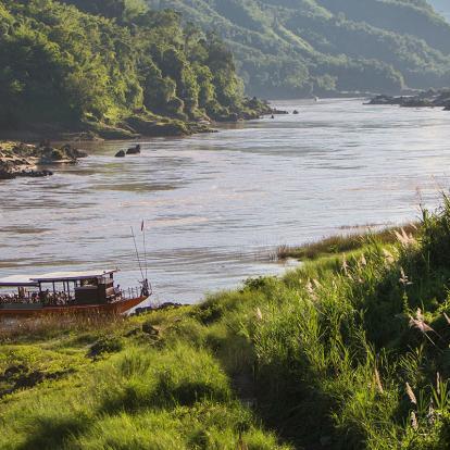 Long, slim boat docking on a river next to forested hills in Pakbeng, Laos