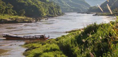 Long, slim boat docking on a river next to forested hills in Pakbeng, Laos