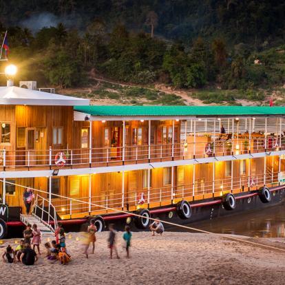 Children stand in front of a wooden traditional cruise boat at dusk in the Laos portion of the Mekong River