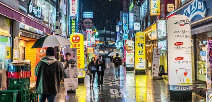 Image of a Korean city in the rain at night, with neon signs and people walkign