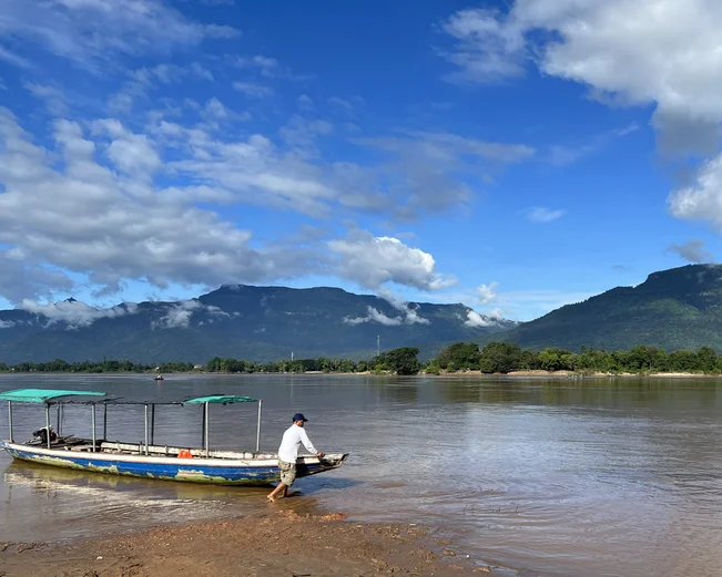Man pulling a long-tail boat ashore on a waterway in Laos, with mountains and clouds in the background.