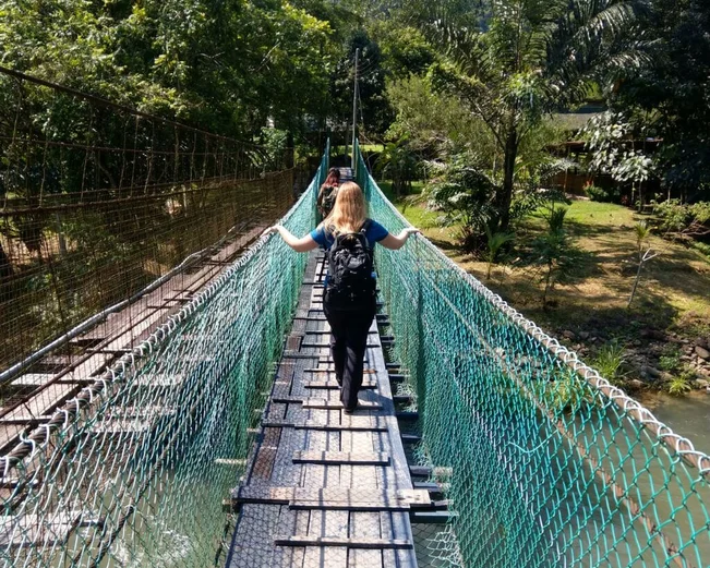Woman crossing a narrow suspension bridge over a river in a tropical forest, wearing a backpack
