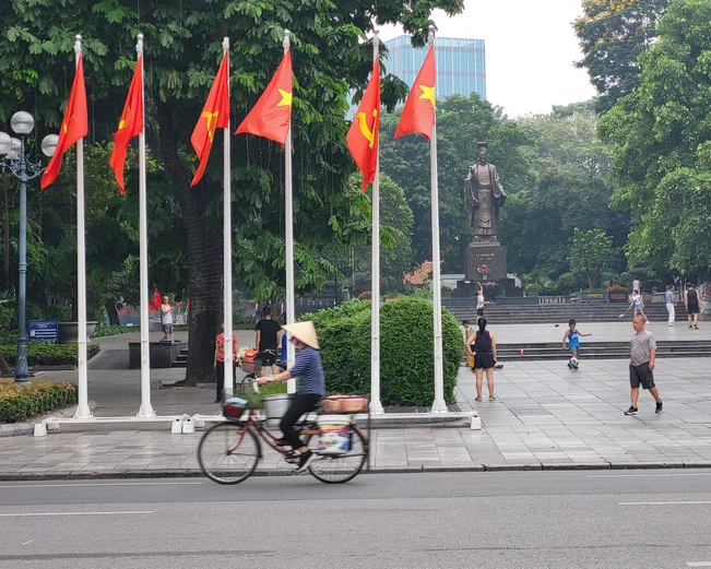 A man wearing a conical hat cycles past a row of Vietnamese flags and a statue of Ho Chi Minh