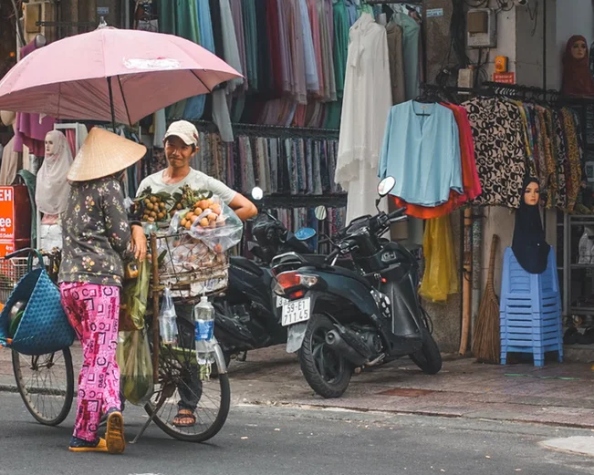 a lady in a conical hat stands with a bike talking to a man outside a clothing store