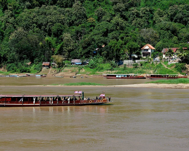A longtail boat cruises past a small riverside village