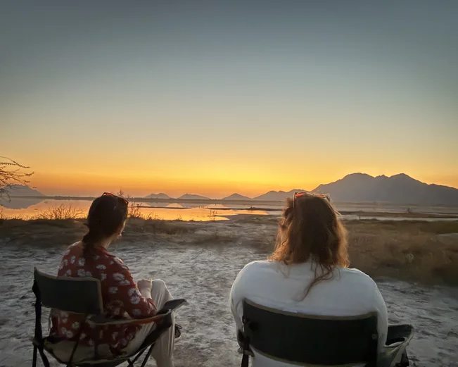Two women in director style chairs watching the sunset, backs to camera.