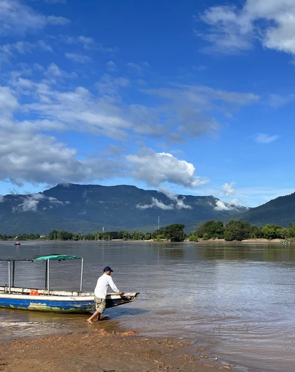 Man pulling a long-tail boat ashore on a waterway in Laos, with mountains and clouds in the background.