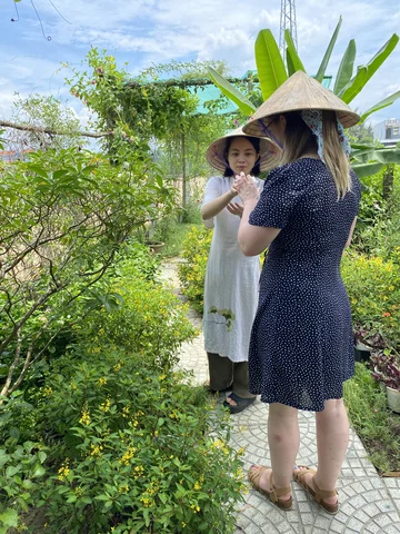 Two people wearing Vietnamese conical hats walking through a tropical garden with green plants, yellow flowers, and banana trees on a sunny day