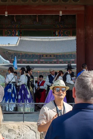 Group of people dressed in traditional Korean hanbok standing under a large wooden gate at a historic palace, with ornate tiled rooftops visible in the background with a tour leader in the foreground