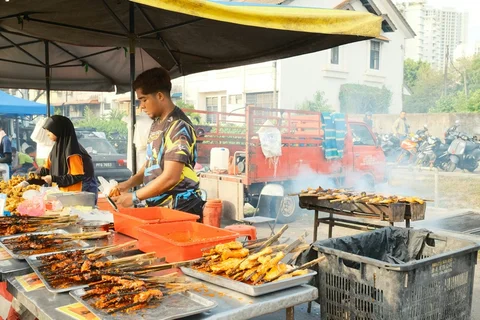 two people man a street food stand, barbecuing various meats on skewers