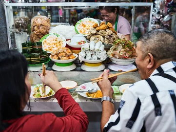 Vietnamese street food stall with plates of fresh herbs, rice noodles, pork rolls, and assorted toppings, as two diners eat with chopsticks at the counter