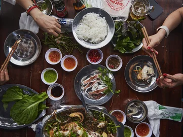 Vietnamese hotpot meal on a wooden table with fresh herbs, rice noodles, dipping sauces, seafood, and vegetables, with diners using chopsticks