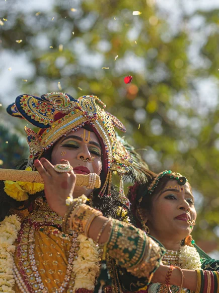 A person dressed in vibrant yellow and gold traditional attire, adorned with an ornate crown, jewelry, and flower garlands, plays a decorated flute while another person stands beside them in green attire and jewelry. Flower petals are falling around them, with a blurred background of trees and sunlight.