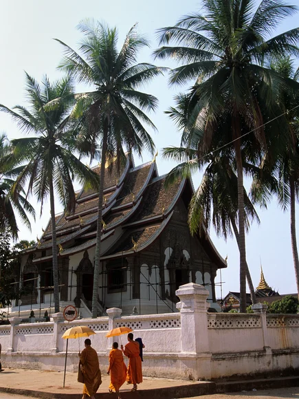 3 monks in traditional wear walking down the street under umbrellas to protect them from the sun past the white temple with pointed roof