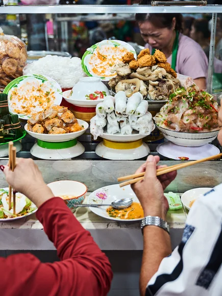Vietnamese street food stall with plates of fresh herbs, rice noodles, pork rolls, and assorted toppings, as two diners eat with chopsticks at the counter