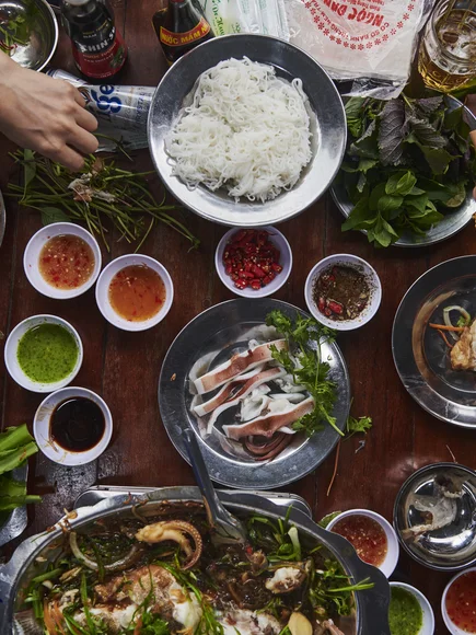 Vietnamese hotpot meal on a wooden table with fresh herbs, rice noodles, dipping sauces, seafood, and vegetables, with diners using chopsticks