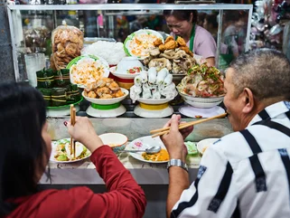 Vietnamese street food stall with plates of fresh herbs, rice noodles, pork rolls, and assorted toppings, as two diners eat with chopsticks at the counter