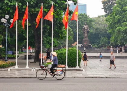 A person in a traditional hat cycling in front of six poles waving the Vietnamese flag with a famous statue in the background