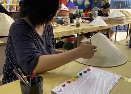 Painting a conical hat, with completed designs hanging on the wall in the background