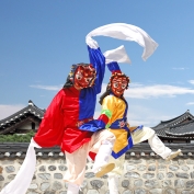 Two traditional folk dancers wearing masks dance in front of hanok houses in South Korea