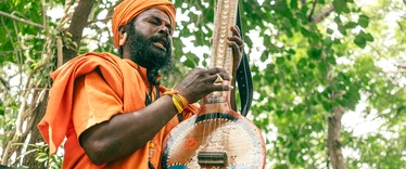 Man playing a stringed instrument. He's wearing an orange turban and robes, with trees in the backdroup