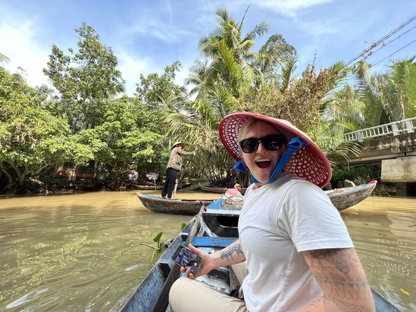 Person sitting in a wooden boat on the Mekong Delta, Vietnam, with palm trees and another boat in the background under a bright blue sky.