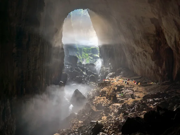 Interior of Son Doong Cave in Vietnam with towering limestone walls, scattered rocks, mist, and sunlight streaming through the cave entrance onto green vegetation.