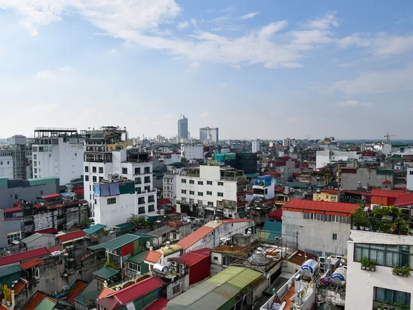 Aerial view of Hanoi, Vietnam showing dense urban buildings with vibrant rooftops and distant skyscrapers under a bright sky.