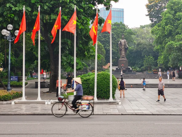 A person in a traditional hat cycling in front of six poles waving the Vietnamese flag with a famous statue in the background