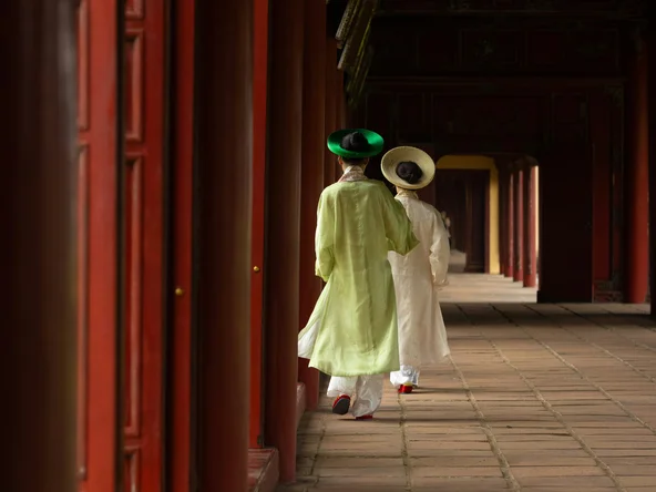 Two Vietnamese women in traditional dress in front of the red doors of the palace in Hue