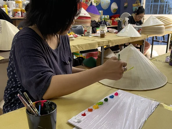 Painting a conical hat, with completed designs hanging on the wall in the background