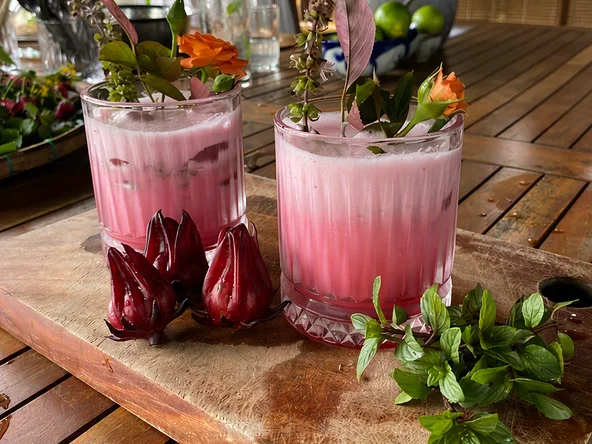 Two pink drinks sit in the forefront. In the background are the ingredients - hibiscus, basil leaves and lemon juice