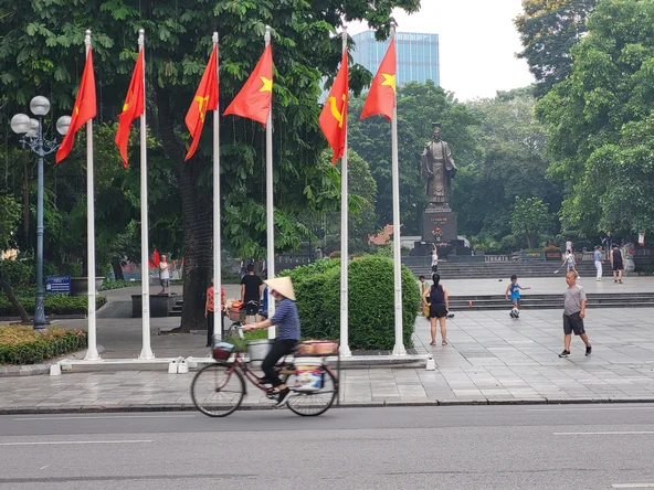 A man wearing a conical hat cycles past a row of Vietnamese flags and a statue of Ho Chi Minh