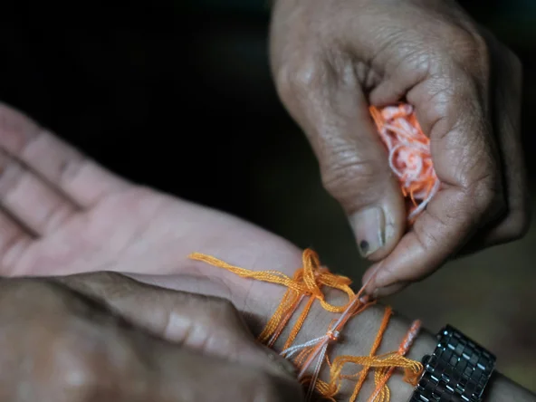 two hands in close up, one tying thin orange strings around the wrist of the other