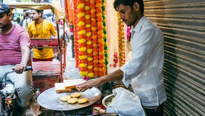Man cooking street food with colourful Mala in the background, tuk tuks are in the street beside him