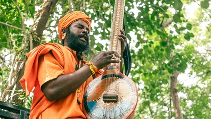 Man playing a stringed instrument. He's wearing an orange turban and robes, with trees in the backdroup