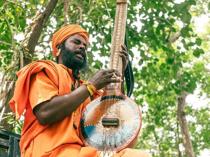 Man playing a stringed instrument. He's wearing an orange turban and robes, with trees in the backdroup
