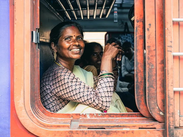 Woman smiling from a train window, the train is red and she's accompanied by fellow passengers inside