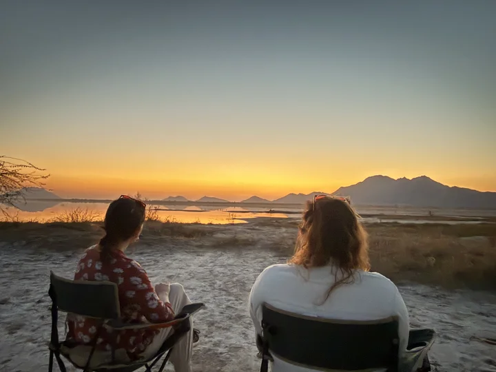 Two women in director style chairs watching the sunset, backs to camera.